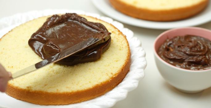 Foto de um bolo aberto ao meio em cima de um prato branco sendo preenchido com creme de chocolate por cima