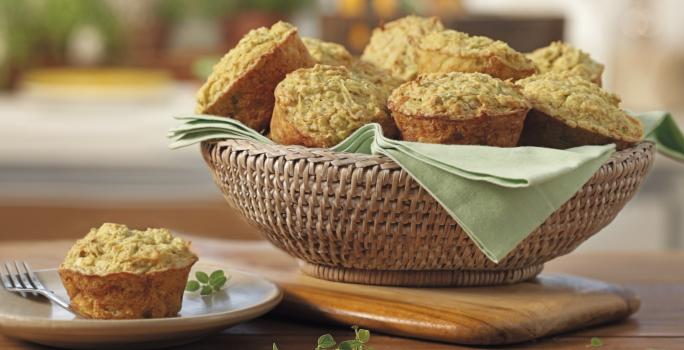 Fotografia em tons de verde em uma bancada de madeira, uma tábua de madeira, uma cesta de vime com vários muffins de aveia e embaixo um paninho verde. Ao lado, um pratinho de sobremesa branco com um muffin em cima dele.