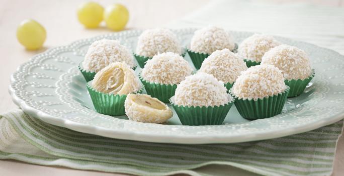 Fotografia em tons de verde e branco de uma bancada vista de frente, contém um pano listrado em verde e branco, um prato redondo branco com vários beijinhos em forma de bolinha com coco por cima.