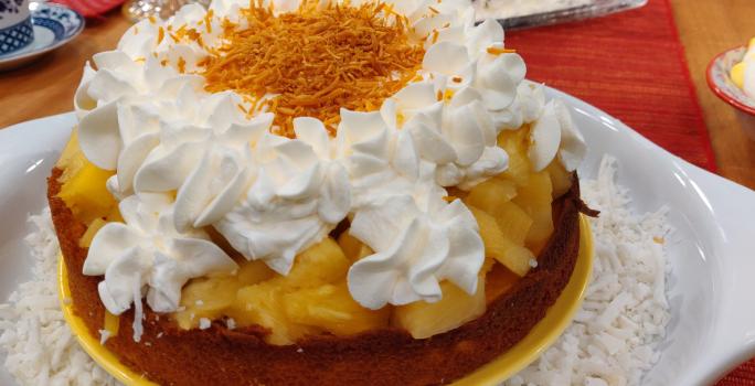 Foto aproximada da receita de bolo de pina colada com leite moça, um doce servido em um prato branco e amarelo, em camadas de massa, abacaxi picado, merengue decorado e coco ralado por cima.