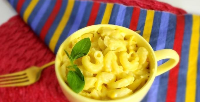 Foto da receita do mac and cheese na caneca servida em uma caneca amarela sobre um pano listrado colorido e ao lado um garfo dourado