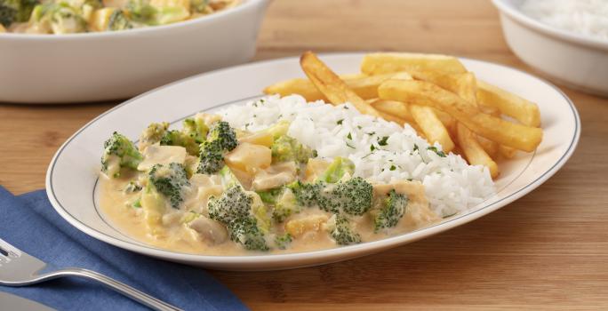 Fotografia em tons de verde em uma bancada de madeira com um guardanapo de pano azul, um prato oval branco no centro com o strogonoff de brócolis, arroz branco e batata frita. Ao fundo, um recipiente com strogonoff de brócolis e outro com arroz.