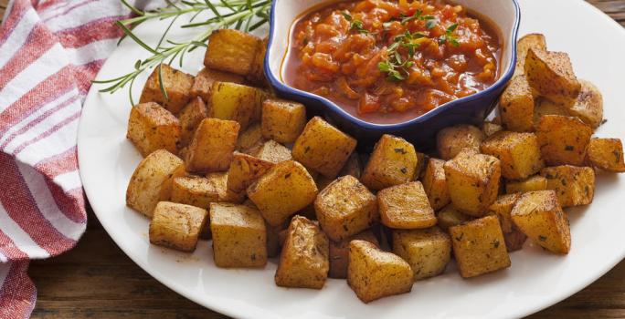 Fotografia em tons de vermelho em uma bancada de madeira escura com um pano branco com listras vermelhas e um prato branco raso grande ao centro com as batatas assadas e um potinho com o molho de tomate picante dentro.