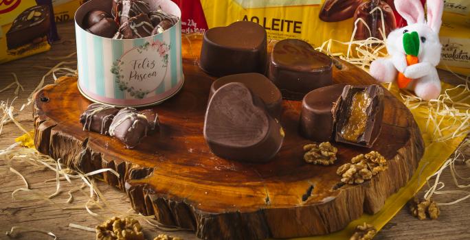 Foto de uma tábua de madeira escura com bombons de chocolate quadrados e em formatos de coração. Um deles está cortado ao meio mostrando o recheio. Tudo está em uma bancada decorada com um tecido amarelo, palha, um coelho de pelúcia e nozes espalhadas