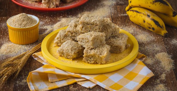 Imagem aproximada de uma mesa de madeira, com uma toalha quadriculada amarela e branca, um prato de madeira amarelo com a receita de palha italiana, algumas bananas inteiras e farinha láctea espalhada pela bancada ao redor