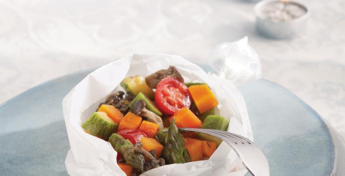 Fotografia em tons de azul em uma mesa com uma toalha branca de renda, um prato branco e azul claro em cima dela e o papillote de legumes (abobrinha, cenoura, tomate) em cima do prato azul, enrolado em um papel manteiga.