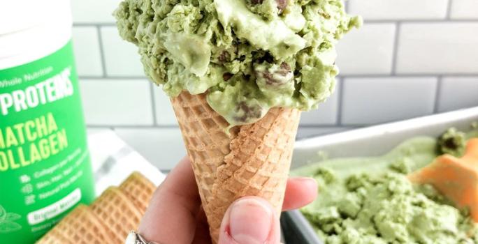 Fotografia em tons de verde em uma bancada de madeira, com um fundo de azulejo, com uma pessoa segurando uma casquinha com o sorvete de matcha, um pote de produto de Vital Proteins. E ao fundo, um recipiente com mais sorvete.