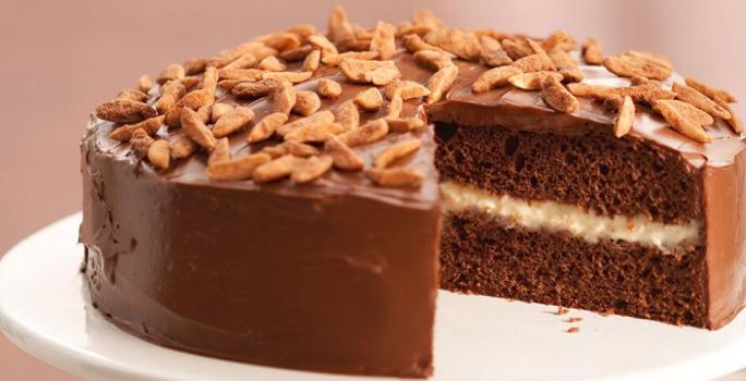 Fotografia em tons de rosa em uma mesa com uma toalha bege, um guardanapo de pano rosinha, um suporte para bolo branco com o bolo de chocolate cortado ao meio em cima dele.