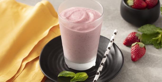 Fotografia em tons de amarelo, branco e preto de uma bancada branca com um prato preto redondo, sobre ele um copo de vidro com smoothie de morango e um canudo branco com bolinhas pretas. Ao lado um paninho laranja e morangos.