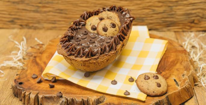 Fotografia em tons de marrom de uma bancada marrom com uma tábua de madeira com um paninho amarelo e branco, sobre ele um ovo recheado de cookie. Ao lado um cookie.
