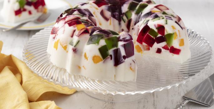 Fotografia em tons de branco e amarelo de uma bancada branca, sobre ele um prato de vidro com a gelatina mosaico. Ao fundo um prato redondo com uma fatia da gelatina. Ao lado um paninho amarelo.