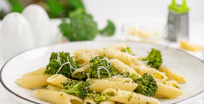 fotografia em tons de branco, bege e verde vista de frente. Contém um prato redondo branco com macarrão tipo penne e brócolis