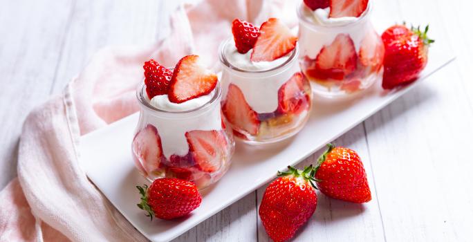 Fotografia de três potinhos de vidro sobre um prato decorativo branco. Dentro dos potes tem um pavê de morango feito com bolo, o creme e coco ralado. Para decorar, muitos pedaços de morango, tanto dentro da sobremesa, quanto na mesa de madeira branca.