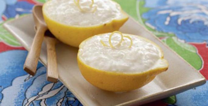 Fotografia em tons de azul, amarelo e marrom de um paninho azul estampado com um prato beje, sobre ela duas metades de um  limão com o arroz doce, uma raspa da limão. Ao lado duas colheres de madeira.