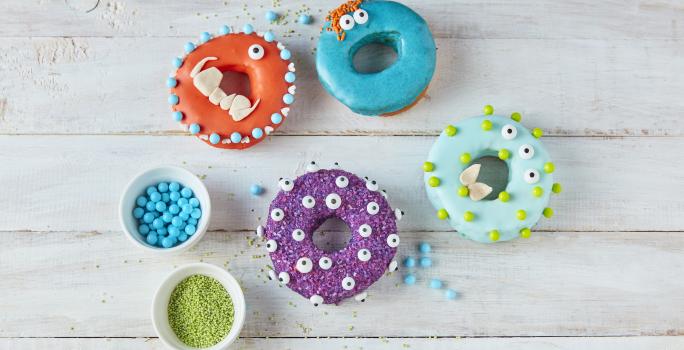 Imagem vista de cima de alguns Donuts Monstro em tons coloridos, decorados com tema de dia das bruxas, tudo em uma bancada de madeira em tom claro