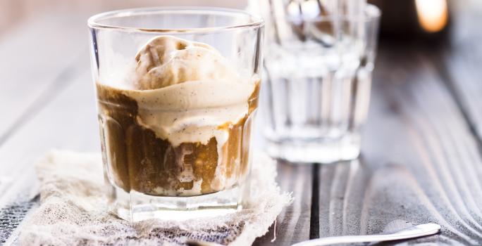 Fotografia de um copo de vidro com café, gelo e sorvete de creme que está em cima de um pano fino, ao lado de uma colher de sobremesa. Ao fundo, um copo de vidro vazio, sobre uma mesa de madeira escura.
