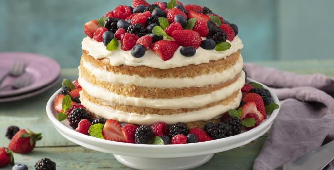 Fotografia em tons de vermelho e lilás em uma bancada de madeira verde, um pano roxo, um suporte para bolo com o bolo pelado (naked cake) com creme branco e com decoração de frutas vermelhas. Ao fundo, pratos pequenos lilás de sobremesa.