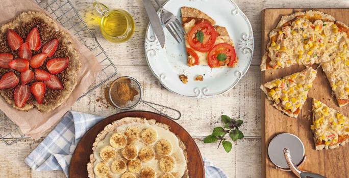 fotografia em tons de bege de uma bancada vista de cima, contém uma tábua redondo e marrom com uma pizza com fatias de banana, uma grade com uma pizza de chocolate e fatias de morango e um prato redondo e branco com uma fatia de pizza