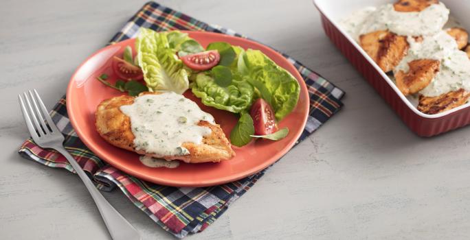 Fotografia em tons de vermelho em uma bancada de madeira cinza com um pano xadrez vermelho, um prato vermelho com o frango com molho de iogurte e ervas e salada dentro dele. Ao lado, um recipiente marrom com mais filés de frango com molho.