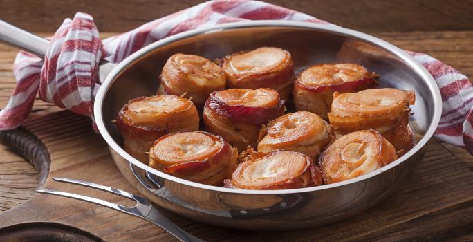 Fotografia em tons de marrom, prata e vermelho de uma bancada de madeira e uma tábua de madeira, sobre ela um panela de inox com medalhões de frango com bacon. Ao lado um garfo de servir, ao fundo um paninho branco e vermelho.