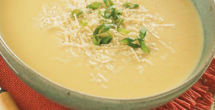 Fotografia em tons de creme e vermelho, com refratário cinza ao centro contendo um creme amarelo decorado com queijo e cebolinha picada sobre guardanapo vermelho, com colher ao lado e tudo sobre aparador de palha na cor bege.