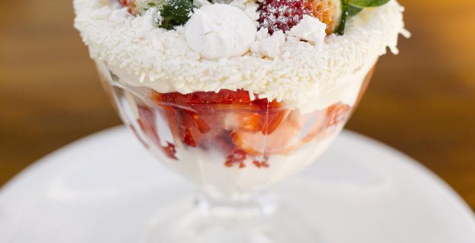 Fotografia de uma taça de sobremesa com merengue, morangos, suspiro e granulado branco nas bordas. O recipiente está em cima de um prato branco de sobremesa, com uma colher pequena. O prato está sobre uma mesa marrom.