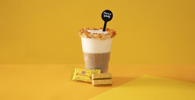 Foto em tons de amarelo da receita de latte salted caramel servida em um copo grande de vidro com um KIT KAT banana ao lado em fundo amarelo