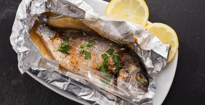 Fotografia em tons de cinza em uma bancada de madeira de cor preta. Ao centro, um prato branco contendo o peixe em um papel alumínio com duas rodelas de limão ao lado.