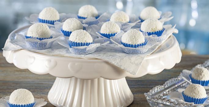 foto em tons de branco e azul de uma bancada cinza vista de frente, contém um suporte para servir bolos branco com diversos docinhos de coco com forminhas azuis