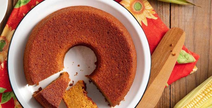 Bolo de milho feito com Farinha Láctea Nestlé em um prato branco, com dois pedaços cortados e espigas de milho em volta.