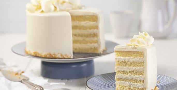 Fotografia em tons de branco, em uma bancada de madeira de cor branca. Ao centro, uma boleira azul contendo o bolo. Ao lado um pires contendo uma fatia de bolo e ao fundo uma jarra branca.