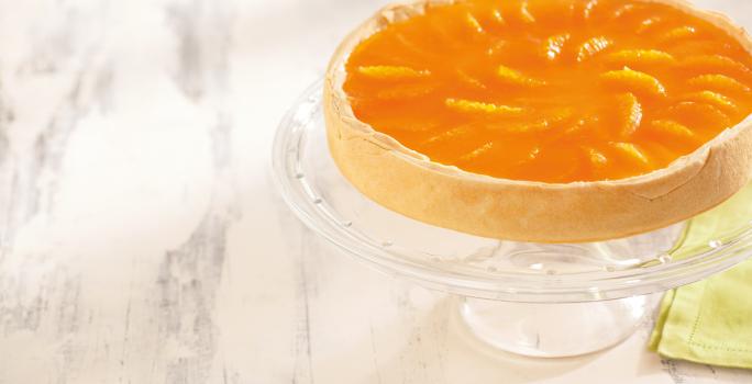 foto em tons de branco e laranja de uma bancada branca vista de cima. Contém um suporte para servir sobremesas transparente com uma torta com cobertura de tangerina por cima.