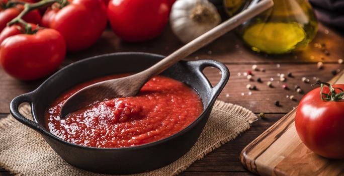 fotografia em tons de marrom e vermelho de uma bancada marrom vista de cima. Contém uma panela preta com molho de tomate e uma colher de madeira para mexer, ao lado esquerdo tomates.