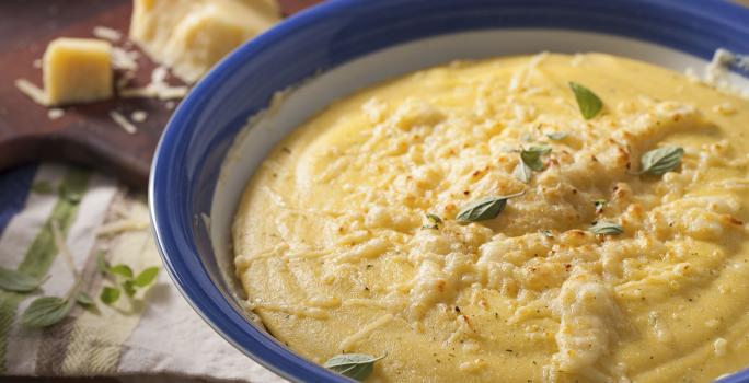 Fotografia em tons de marrom e azul de uma bancada marrom com um paninho branco, sobre ele um recipiente redondo branco e azul com polenta. Ao fundo uma tábua com queijo parmesão.