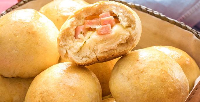 Fotografia em tons de amarelo em uma bancada de madeira vermelha um pano azul, uma cesta de vime com pães recheados com queijo.