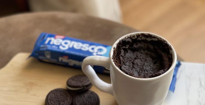 Foto de uma caneca branca contendo a receita de Bolo Negresco, sobre uma tábua com alguns Biscoitos Negresco soltos e um pacote em tom azul de Biscoito Negresco