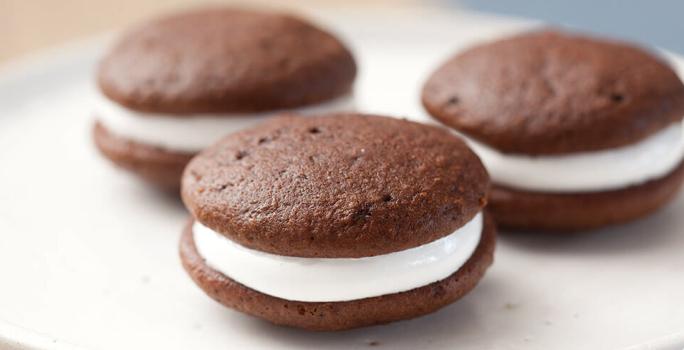 Fotografia em tons de preto em uma bancada de madeira clara, um prato redondo branco raso com três sanduicinhos de chocolate recheados com marshmallow.