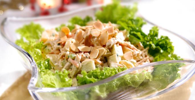 Fotografia em tons de verde em uma mesa com uma toalha branca, um apoiador de prato de madeira, um recipiente de vidro fundo com a salada de alface e frango dentro dele.