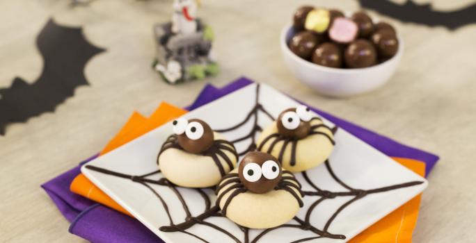 Imagem aproximada de um prato quadrado branco com três biscoitos em formatos de aranhas. A bancada está decora com papéis em tons roxo e laranja e alguns morcegos de mentira.
