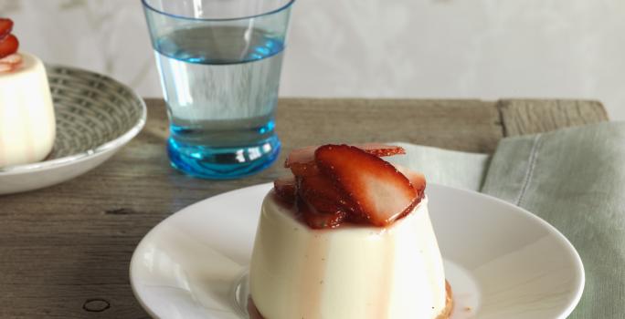 Fotografia de um flan de canela com calda de morangos em um prato branco fundo sobre uma mesa de madeira. Ao fundo, outro flan de canela e, ao lado, um copo d'água.