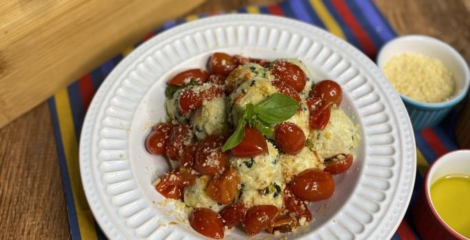 Fotografia em tons de amarelo e vermelho em uma bancada de madeira com um pano colorido em azul, vermelho e amarelo e um prato branco fundo em cima dele com o gnudi e molho de tomate por cima dentro do prato. Ao lado, potinhos com queijo ralado e azeite.