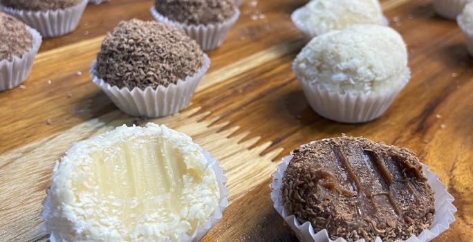 Fotografia em tons de marrom e branco em uma bancada de madeira de cor marrom. Ao centro, estão várias forminhas enfileiradas com brigadeiros de chocolate e brigadeiro branco.