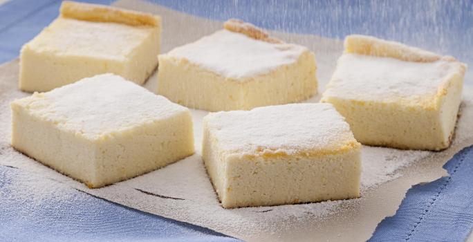 Fotografia em tons de amarelo, azul e branco de uma bancada branca com paninho azul, sobre ele pedaços de torta de ricota polvilhadas com açúcar.
