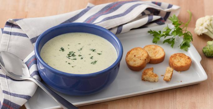 Fotografia em tons de azul e verde, com cumbuca azul ao centro com creme de legumes e salsa picada com torradinhas sobre travessa branca, ramo de salsa, brócolis e couve-flor decorando, guardanapo branco e azul com colher, sobre bancada de cor palha.