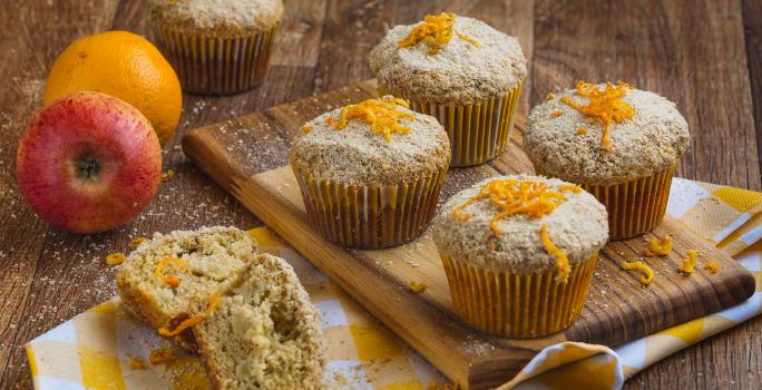 Imagem aproximada de muffins em tom claro, sobre uma tábua de madeira em um pano quadriculado em tom amarelo e branco. Ao fundo há uma maçã e uma laranja e alguns farelos de Farinha Láctea.