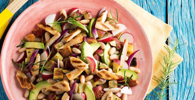 Fotografia em tons de azul e rosa em uma bancada de madeira azul com um prato rosa com salada de batata-doce, frango, feijão branco e abacate dentro dele.