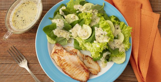 Fotografia em tons de verde em uma bancada de maneira de cor marrom. No centro, um prato redondo azul contendo a salada e um filé de peixe grelhado. Ao fundo, um pano laranja, um garfo e um recipiente com molho.