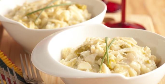 foto em tons de marrom, branco e vermelho tirada de uma bancada vista de frente, contém dois recipientes redondos brancos com o risoto com aspargos. Ao fundo duas taças vermelhas e à frente dois garfos para servir.