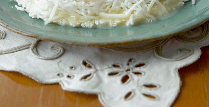 Foto com uma tapioca de Leite Moça e coco em um prato na cor azul, em cima de uma toalha branca decorada