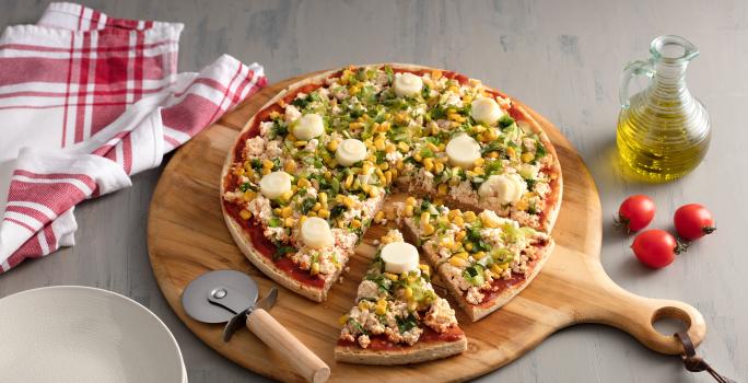 Fotografia em tons de vermelho em uma bancada de madeira cinza com uma tábua de pizza de madeira com a pizza em cima decorada com escarola e ricota. Ao lado, um vidro de azeite, tomates e um cortador de pizza.